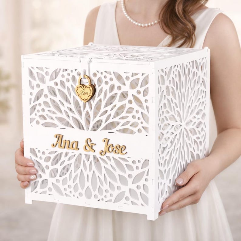May include: A white, laser-cut wooden card box with floral patterns. The box features the names "Ana & Jose" in gold lettering and a wooden heart-shaped lock. The box is held by a person wearing a white dress.