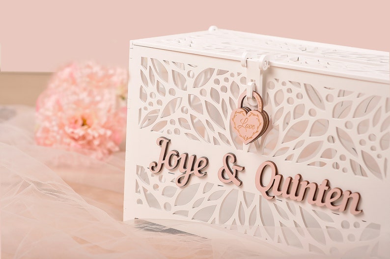 May include: A white, laser-cut wooden card box with a floral design. The box features the names "Joye & Quinten" in pink script. A heart-shaped lock with the word "Love" secures the lid. Soft pink flowers and fabric are in the background.