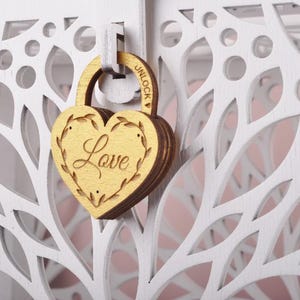 May include: A wooden heart-shaped padlock with the word "Love" engraved on it, hanging on a white, laser-cut wooden structure. The padlock is gold-colored, and the structure has a floral design. The word "UNLOCK" is visible on the padlock.