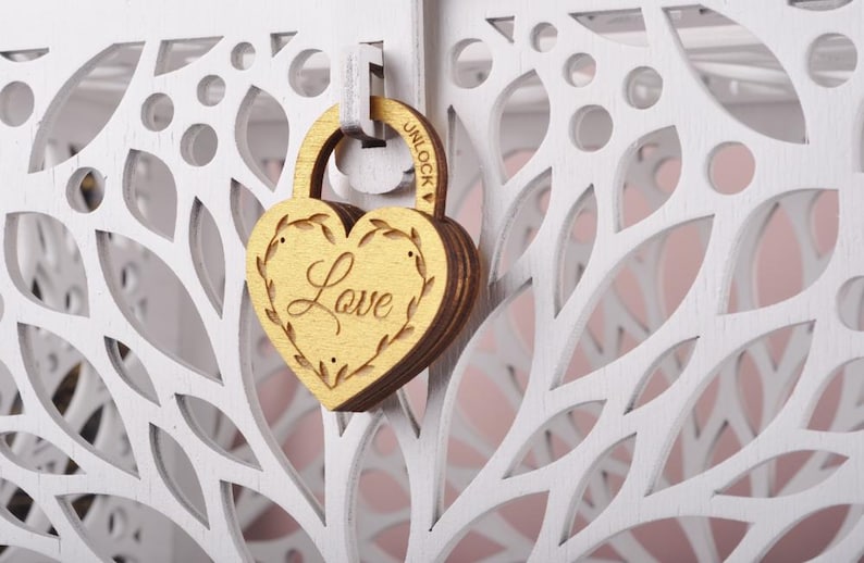 May include: A wooden heart-shaped padlock with the word "Love" engraved on it, hanging on a white, laser-cut wooden structure. The padlock is gold-colored, and the structure has a floral design. The word "UNLOCK" is visible on the padlock.