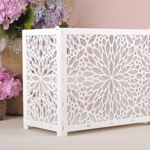 May include: A white, rectangular decorative screen with a floral cut-out design. The screen is made of wood and has a folding design. The screen is placed in front of a pink background with flowers.