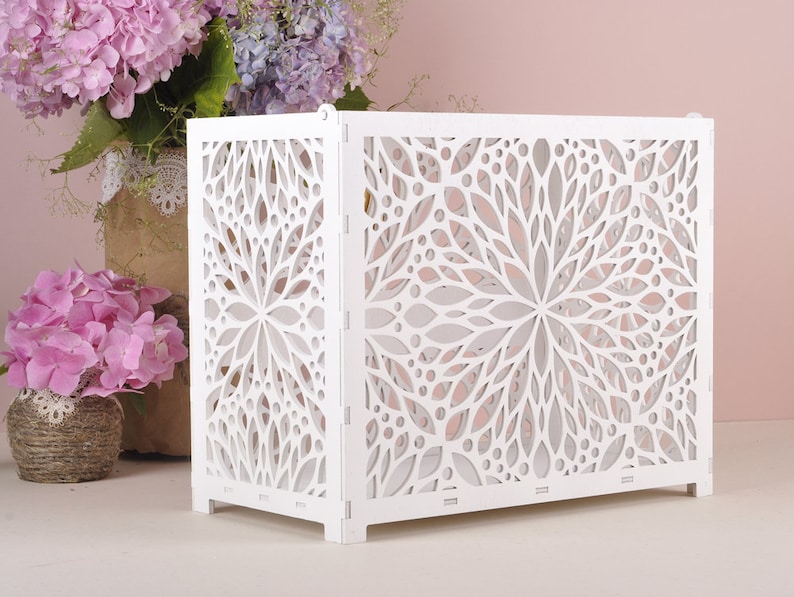 May include: A white, rectangular decorative screen with a floral cut-out design. The screen is made of wood and has a folding design. The screen is placed in front of a pink background with flowers.