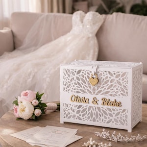 May include: A white wooden card box with a floral design and the names "Olivia & Blake" in gold lettering. The box has a heart-shaped lock. A bouquet of pink and white peonies and a wedding dress are in the background.