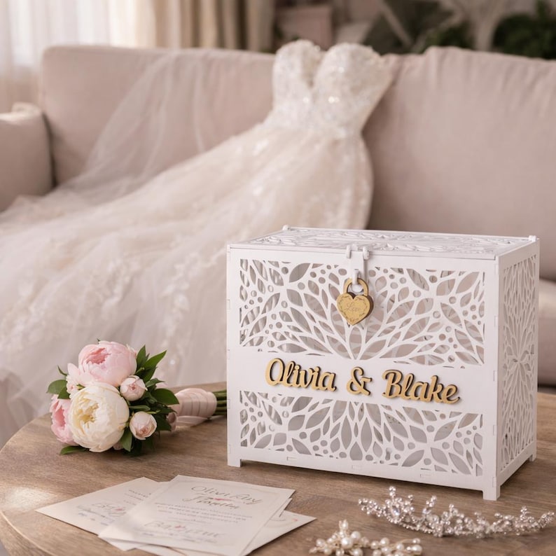 May include: A white wooden card box with a floral design and the names "Olivia & Blake" in gold lettering. The box has a heart-shaped lock. A bouquet of pink and white peonies and a wedding dress are in the background.