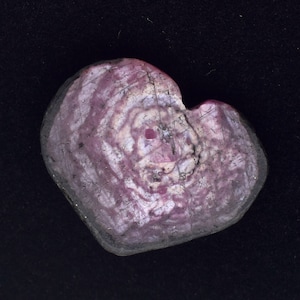 Può includere: Una pietra a forma di cuore con un motivo a spirale di tonalità rosa e bianche. La pietra ha un bordo grigio scuro e una piccola inclusione rossastra vicino al centro. Lo sfondo è nero pieno.