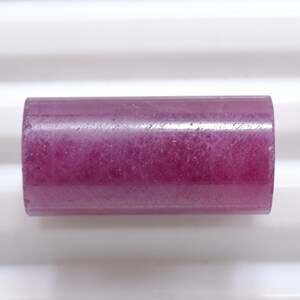 Op de afbeelding: Een cilindrische, gepolijste edelsteen in een diepe magenta tint. De edelsteen heeft een gladde, licht doorschijnende uitstraling. De vorm is een korte cilinder, met een consistente diameter over de lengte. De edelsteen staat tegen een witte achtergrond.
