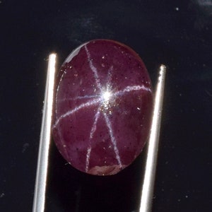 May include: A deep purple star ruby gemstone with a six-rayed star in the center. The stone is oval shaped and is held in place by two silver tweezers.