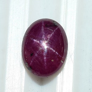 May include: Oval-shaped, deep red gemstone with a six-rayed star pattern. The polished surface of the ruby reflects light, creating a star effect. The gemstone is set against a white background.