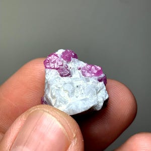 Könnte beinhalten: Ein rohes Mineralexemplar mit leuchtend magentafarbenen kristallinen Strukturen, die in eine raue, undurchsichtige weiße Matrix eingebettet sind. Die Kristallformationen weisen ausgeprägte geometrische Facetten und eine durchscheinende Qualität auf.