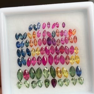 Könnte beinhalten: Eine Sammlung loser, facettierter Edelsteine in einer weißen Präsentationsbox. Die Steine sind überwiegend im Marquise-Schliff gehalten und in verschiedenen Farben wie Blau, Pink, Gelb, Grün und Violett, mit einer Reihe kleinerer Tropfensteine.