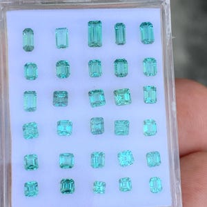 K&ouml;nnte beinhalten: Eine Sammlung von 28 Smaragdschliff-Edelsteinen in einer durchsichtigen Plastikbox. Die Edelsteine sind in verschiedenen Gr&uuml;nt&ouml;nen und Blaut&ouml;nen.
