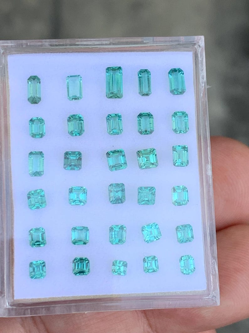 K&ouml;nnte beinhalten: Eine Sammlung von 28 Smaragdschliff-Edelsteinen in einer durchsichtigen Plastikbox. Die Edelsteine sind in verschiedenen Gr&uuml;nt&ouml;nen und Blaut&ouml;nen.