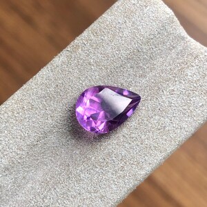 Könnte beinhalten: Ein tropfenförmiger Amethyst-Edelstein mit tiefem Lilafarbton. Der Edelstein ist facettiert und hochglanzpoliert.