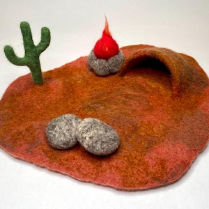 May include: A felt play set with a red campfire, a green cactus, two gray rocks, and a brown cave opening. The set is on a red, orange, and brown felt background.