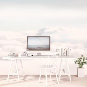 Puede incluir: Un escritorio blanco con un monitor de computadora que muestra un paisaje marino, una silla blanca, una pila de libros, una taza, gafas y auriculares. La pared detrás del escritorio presenta un mural de nubes blancas sobre un cielo pálido.