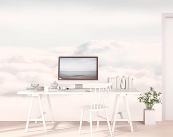 May include: A white desk with a computer monitor displaying a seascape, a white chair, a stack of books, a mug, glasses, and headphones. The wall behind the desk features a large-scale mural of soft, white clouds against a pale sky.