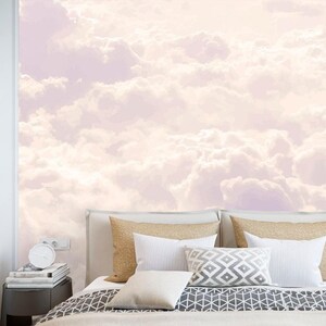 Puede incluir: Un mural de pared que presenta una representación suave y pictórica de nubes esponjosas en tonos rosa pálido, crema y lavanda claro. El mural cubre la pared detrás de una cama con almohadas de color beige, blanco y gris y una colcha geométrica gris.