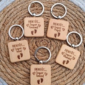 Peut inclure: Porte-clés en bois avec anneaux argentés. Chaque porte-clés porte les mots "BIENTÔT... Super" suivis du titre d'un membre de la famille et d'empreintes de pas de bébé. Les porte-clés sont disposés sur un tapis circulaire tissé.