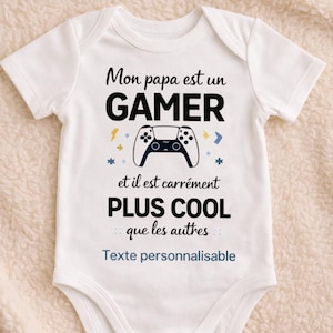 Könnte beinhalten: Weißer Baby-Body mit dem Text "Mon papa est un GAMER" und "et il est carrément PLUS COOL que les autres". Das Design beinhaltet einen schwarzen Gamecontroller und blaue und gelbe Akzente. Auf dem Boden des Bodys steht "Texte personnalisable".