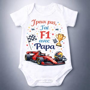 Może przedstawiać: Białe body dla dziecka z grafiką czerwonego samochodu Formuły 1. Tekst brzmi "J'peux pas, J'ai F1 avec Papa" w kolorze czerwonym i niebieskim. Zawiera flagę w szachownicę, trofeum i kask wyścigowy.