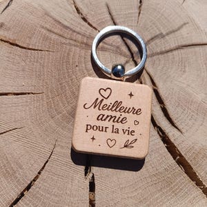 Peut inclure: Un porte-clés en bois avec un anneau argenté et une étiquette carrée. L'étiquette est gravée des mots "Meilleure amie pour la vie" en écriture cursive, ainsi que des motifs de cœur, d'étoile et de feuille. Le porte-clés est posé sur un fond en bois.