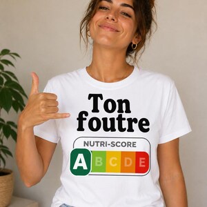 Puede incluir: Camiseta blanca con el texto "Ton foutre" en negro, encima de una etiqueta Nutri-Score con las notas A a E. La persona lleva vaqueros azules y hace un gesto con la mano.