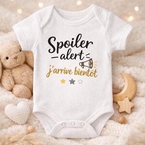 Puede incluir: Body de bebé blanco con el texto "Spoiler alert - j'arrive bientôt" en negro y dorado. El body presenta un gráfico de megáfono y detalles de estrellas. Un osito de peluche y una luna y estrella de madera están en el fondo.