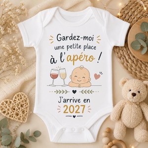 Peut inclure: Un body pour bébé blanc à manches courtes avec le texte 'Gardez-moi une petite place à l'apéro !' en noir et doré, accompagné d'illustrations de deux verres à vin, d'un visage de bébé souriant, d'un cœur et du texte 'J'arrive en 2027'.