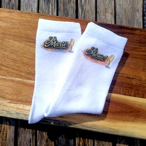 May include: A pair of white socks with the words "Le Marie" and "Mariée!" in a script font, along with a champagne glass graphic. The socks are displayed on a wooden surface.