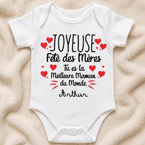 Peut inclure: Body de bébé blanc avec l'inscription "JOYEUSE Fête des Mères Tu es la Meilleure Maman du Monde Arthur" en noir. Des cœurs rouges ornent le texte. Le body a des manches courtes et une fermeture à boutons-pression.