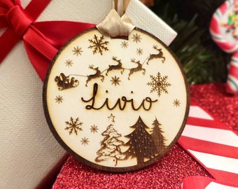 Peut inclure: Ornement de Noël rond en bois avec le nom "Livio" gravé en cursive. Il présente des images découpées au laser du traîneau du Père Noël, des rennes, des flocons de neige et des sapins. Suspendu par un ruban beige.