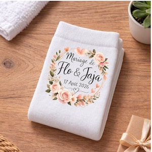 May include: White folded towel with a floral wreath design. The wreath surrounds the text "Mariage de Flo & Jaja" and the date "17 Août 2026". The design includes peach-colored roses and green leaves.