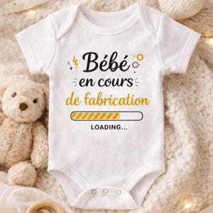 Könnte beinhalten: Weißer Baby-Body mit dem französischen Text "Bébé en cours de fabrication" in Schwarz und Gold. Das Design beinhaltet eine Ladebalken-Grafik. Ein Teddybär befindet sich neben dem Body.