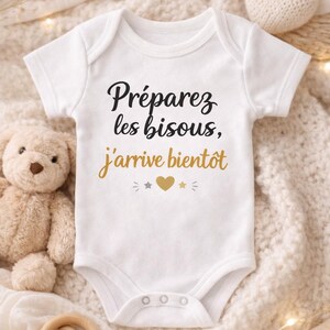 Op de afbeelding: Witte babybody met de Franse tekst "Préparez les bisous, j'arrive bientôt" in zwart en goud. De body heeft een drukknoopsluiting en is omgeven door een teddybeer, een houten speelgoed en een gebreid hart.