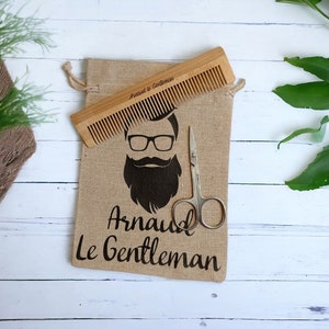 May include: A burlap bag with a drawstring closure, featuring a black and white illustration of a man with a beard and glasses. The bag contains a wooden comb and a pair of small scissors. The text "Arnaud Le Gentleman" is printed on the bag.