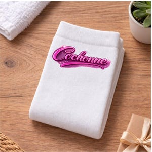 May include: A stack of folded white towels with the word "Cochonne" in pink script. The towels are on a wooden surface, with a small plant and a wrapped gift in the background.