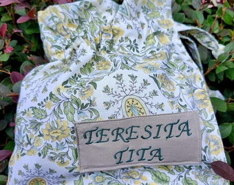 May include: A white fabric drawstring bag with a floral pattern and a brown label that says "TERESITA TITA".