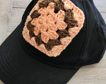 May include: A black baseball cap with a square, crocheted patch. The patch features a floral design in shades of peach and brown. The cap has a curved brim and a button on top.