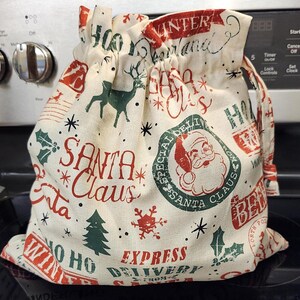 May include: A drawstring gift bag with a festive Christmas theme. The bag is cream-colored with red and green illustrations of Santa Claus, reindeer, snowflakes, and text that reads "Santa Claus" and "Express Delivery".