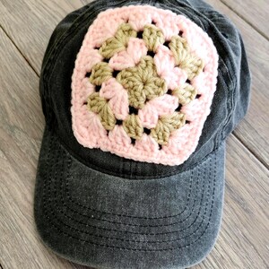 May include: A dark gray baseball cap with a crocheted square design. The square features a pink and tan granny square pattern, adding a handmade touch to the cap. The cap has a curved brim and a classic, casual style.