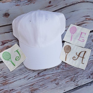 May include: White baseball cap with a green tennis racket and the letter J embroidered on a white patch. There are two other patches with a pink tennis racket and the letters E and A embroidered on them.