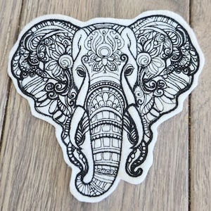 Könnte beinhalten: Ein weißer Aufnäher mit einem detaillierten schwarzen Elefanten-Design. Der Elefantenkopf ist zentriert und zeigt komplizierte Muster und florale Motive. Der Aufnäher hat ein weiches, strukturiertes Aussehen und eignet sich für verschiedene Anwendungen.