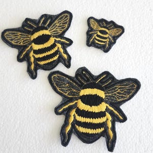 May include: Three embroidered bee patches in varying sizes. The bees feature black and yellow striped bodies, black wings with gold detailing, and are outlined in black. These patches are suitable for clothing or craft projects.