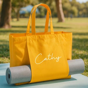 May include: A mustard yellow tote bag with the name "Cathy" in white script. A gray yoga mat is rolled up and secured inside the bag. The bag is sitting on a light blue yoga mat, outdoors.