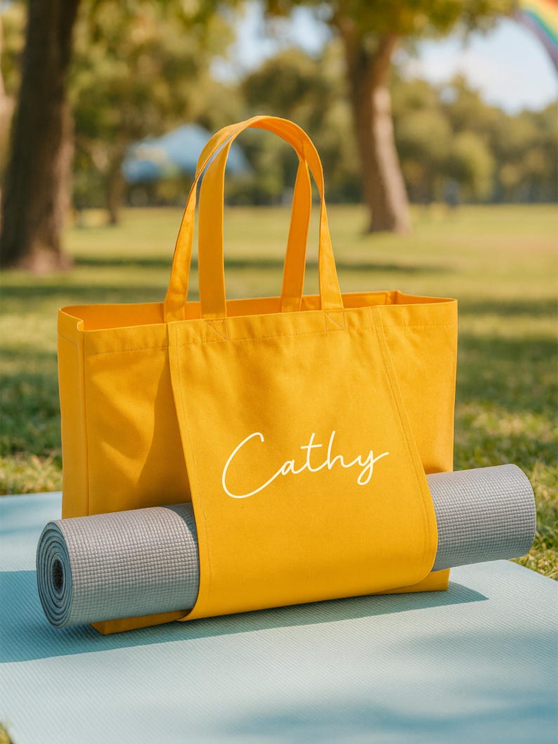May include: A mustard yellow tote bag with the name "Cathy" in white script. A gray yoga mat is rolled up and secured inside the bag. The bag is sitting on a light blue yoga mat, outdoors.