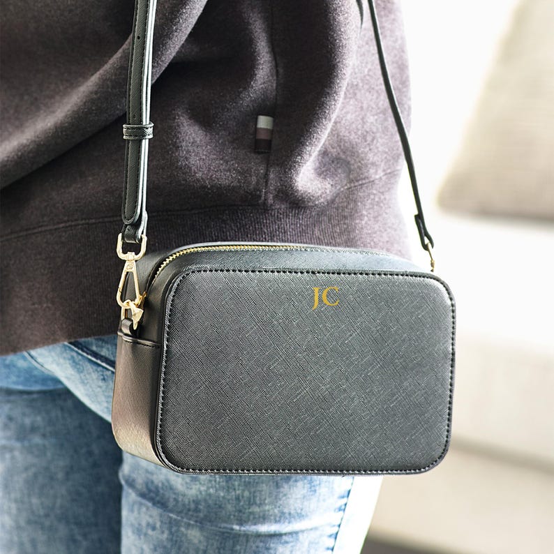 May include: A black crossbody bag with a gold zipper and strap hardware. The bag has a textured surface and the initials "JC" in gold lettering. The bag is rectangular and has a long, thin strap.