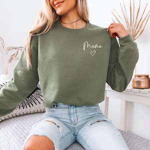 May include: Olive green sweatshirt with the word "Mama" in white script and a small heart. The sweatshirt is a crew neck style and has long sleeves. The model is wearing light blue jeans.
