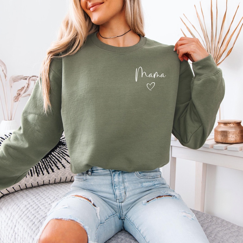 May include: Olive green sweatshirt with the word "Mama" in white script and a small heart. The sweatshirt is a crew neck style and has long sleeves. The model is wearing light blue jeans.