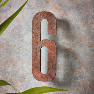 May include: A rusty metal number six, with a rough, textured surface. The number is attached to a wall with a light brown, textured surface.