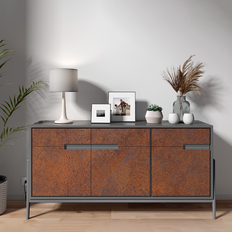 K&ouml;nnte beinhalten: Ein rechteckiges Sideboard mit einem dunkelgrauen Rahmen und drei Schrankt&uuml;ren in rostfarbener, texturierter Optik. Auf der Oberseite stehen eine wei&szlig;e Tischlampe, zwei gerahmte Fotos, eine kleine Sukkulente und eine Glasvase mit getrocknetem Gras.